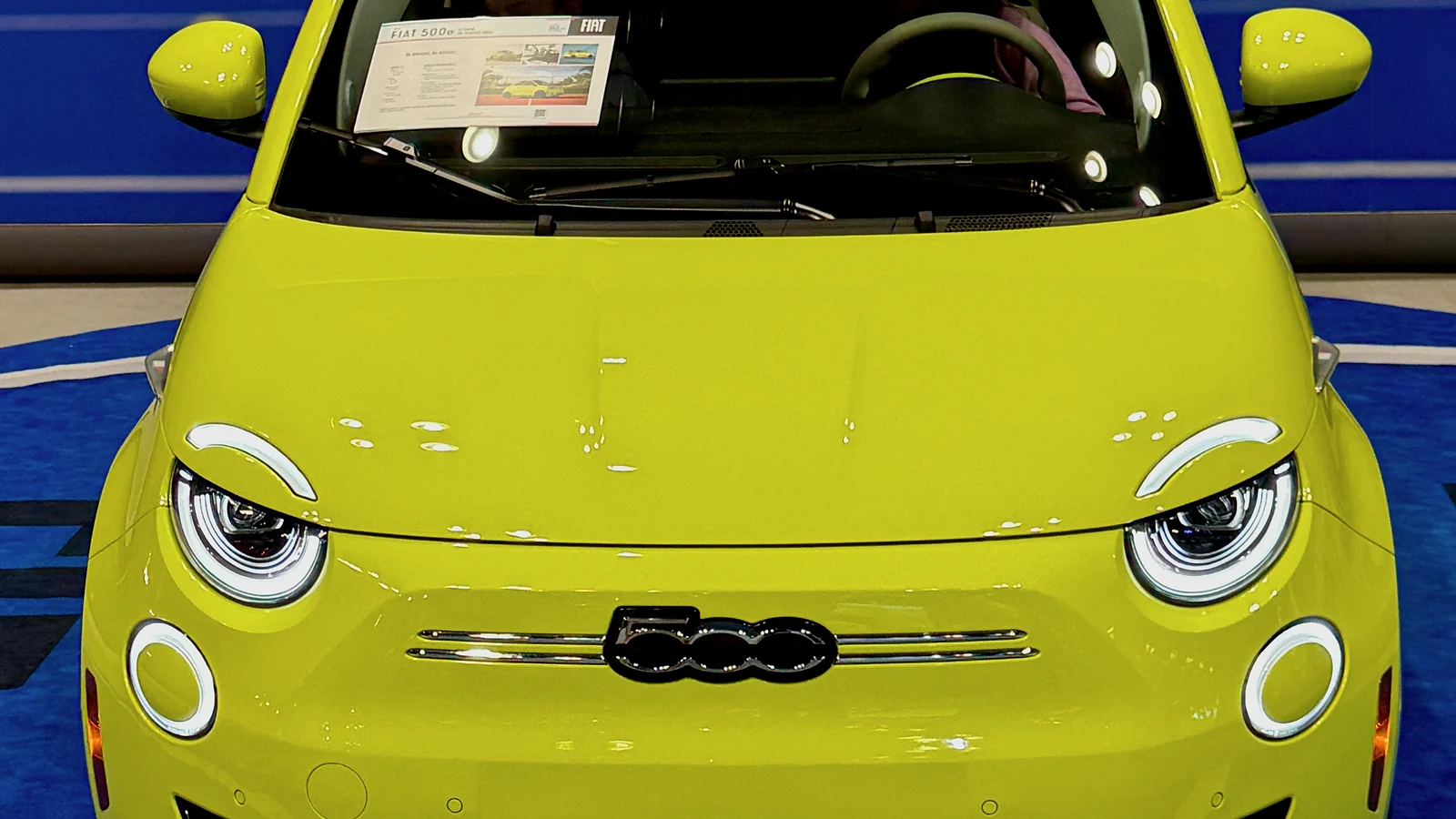 Close frontal view of a bright yellow 2025 Fiat 500e electric hatchback displayed under motor-show lights