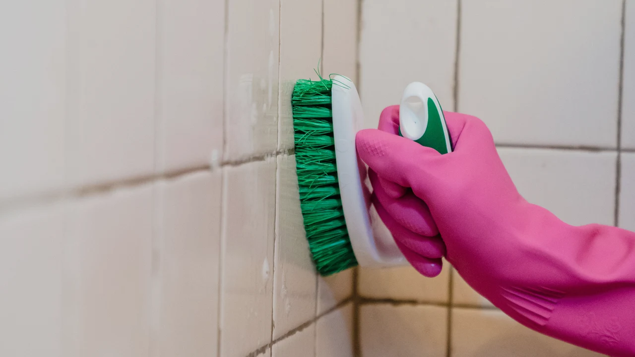 A pink-gloved hand using a small green brush along the grout between bathroom tiles