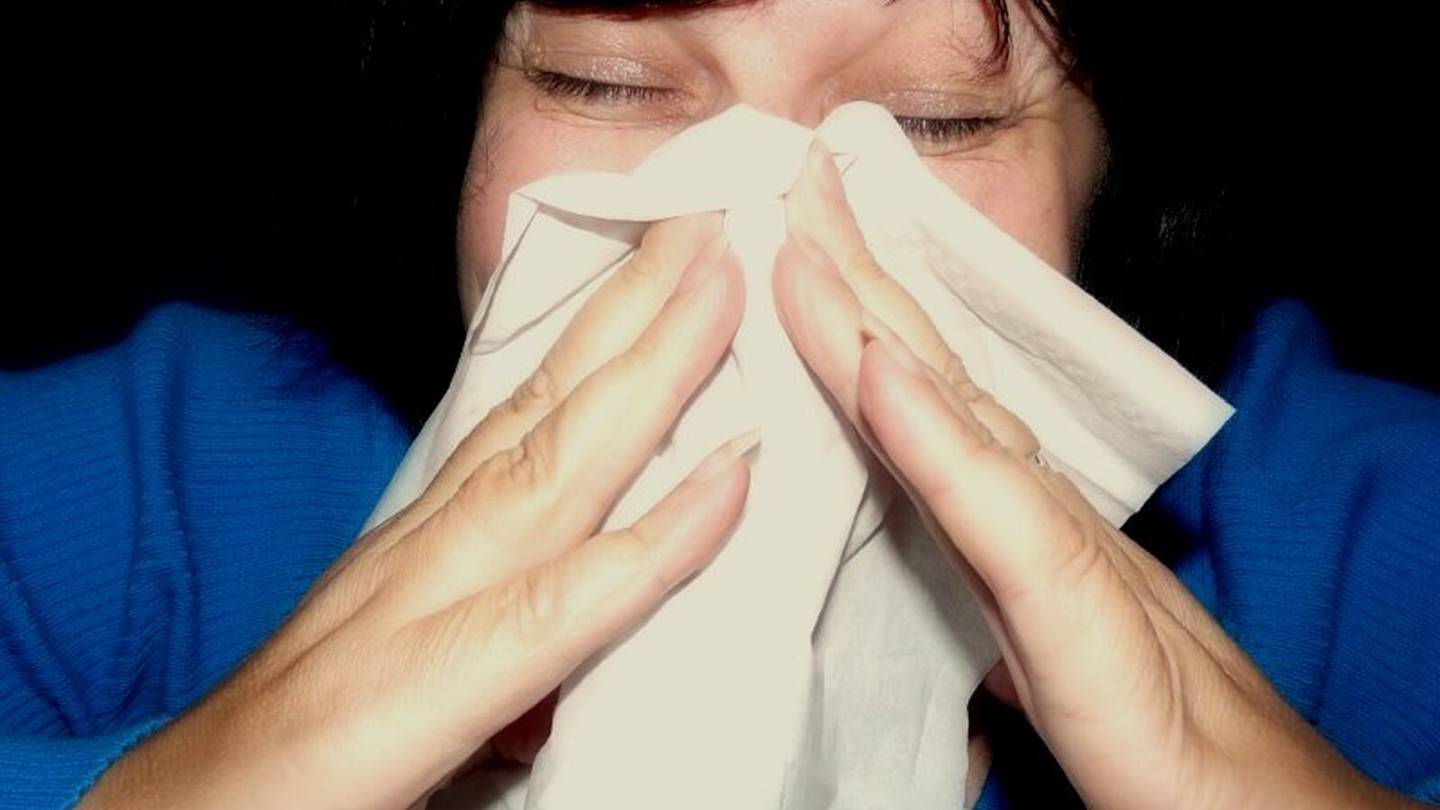 A person covering an involuntary sneeze with a white handkerchief against a dark background