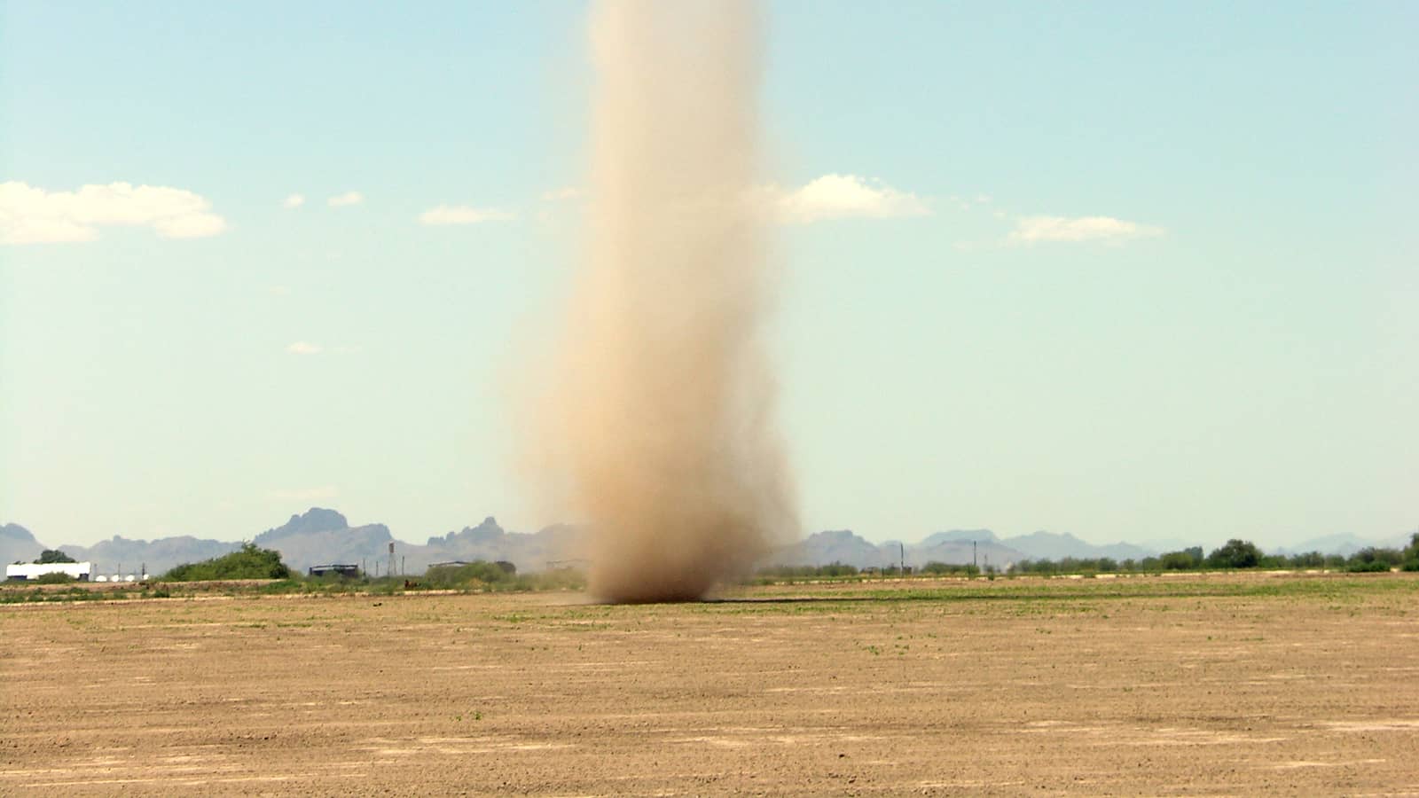 A tall dust devil rising from a dry field beneath a mostly clear blue sky in Arizona